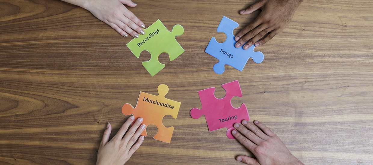 Four individual hands are holding four different colored puzzle pieces with the words recordings, songs, merchandise, and touring printed on them